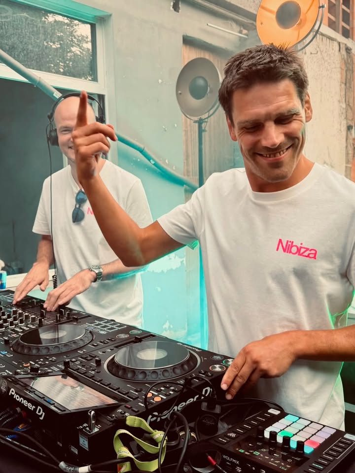 Twee dj's lachend achter de decks, een van hen met een vinger in de lucht.