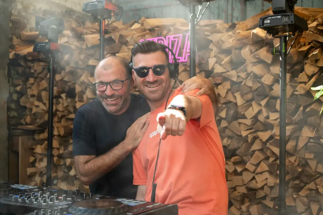 Twee dj's poseren voor een wand van gestapeld haardhout, met het neonbord erachter.